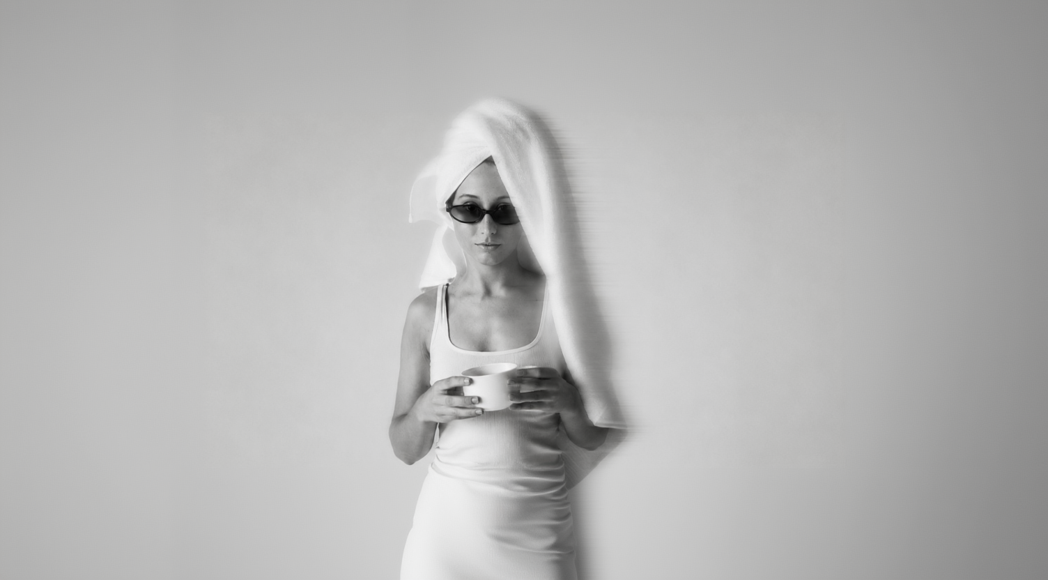 A black and white lifestyle photograph of a woman with a white towel wrapped around her head, wearing dark sunglasses and a white tank top while holding a white mug.
